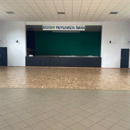 PARKIETY VIP - Sala z parkietem ułożonym we wzór jodełki, kontrastującym z szarymi płytkami. Scena z zielonym tłem i napisem 'Jesteśmy Przyszłością Świata' nad nią.