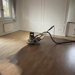 PARKIETY VIP - Cyklinowanie parkietu w pokoju: połowa podłogi wycyklinowana, ukazując jaśniejszy odcień drewna, maszyna do cyklinowania w trakcie pracy, eleganckie białe ściany.