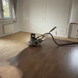Cyklinowanie parkietu w pokoju: połowa podłogi wycyklinowana, ukazując jaśniejszy odcień drewna, maszyna do cyklinowania w trakcie pracy, eleganckie białe ściany.