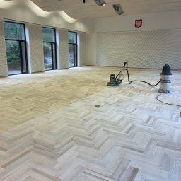 PARKIETY VIP - Sala z jasnym parkietem w jodełkę, częściowo wyszlifowanym, z maszyną do cyklinowania i odkurzaczem przemysłowym. W tle okna i godło Polski.
