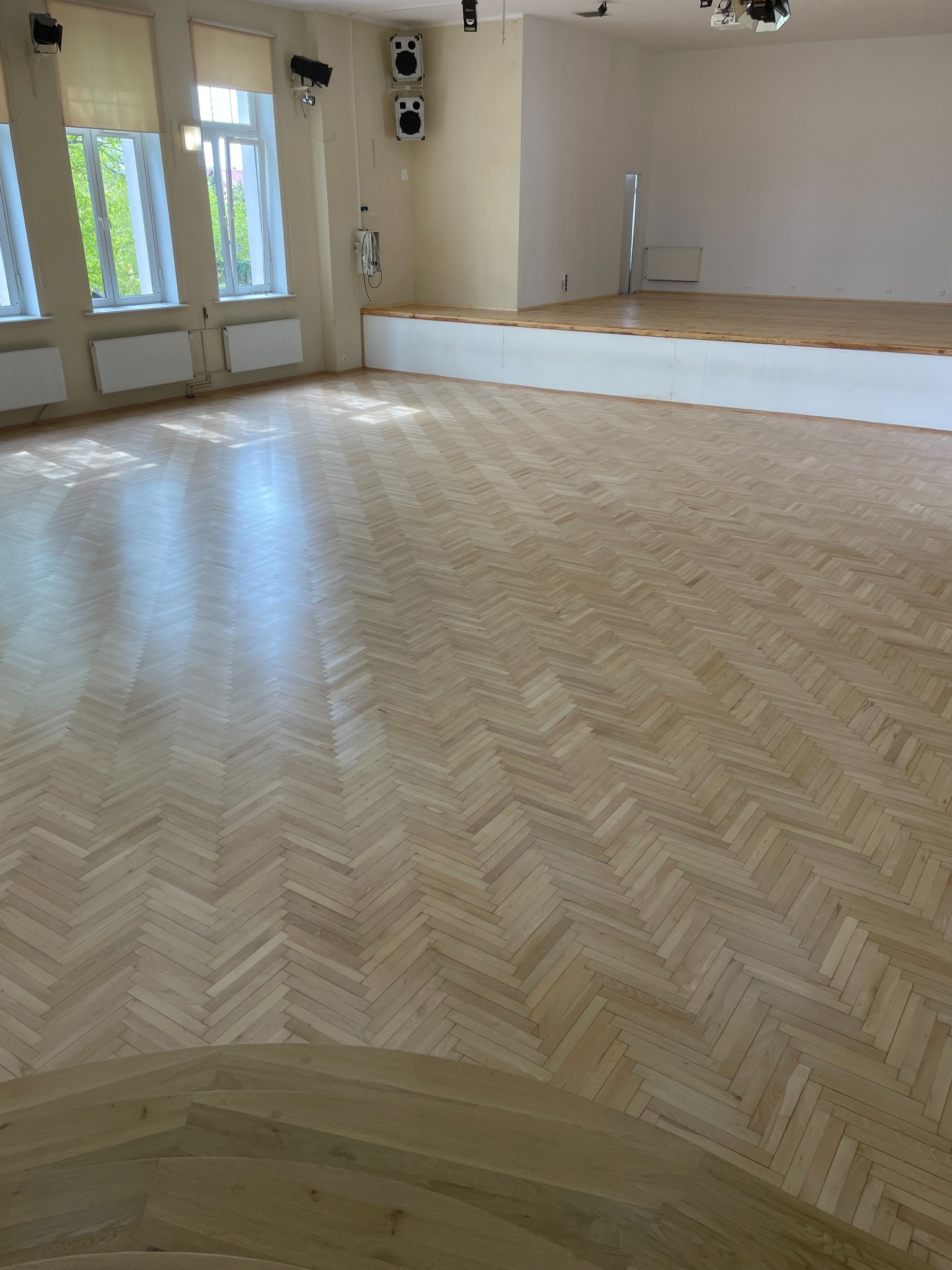 Jasna, nowa podłoga parkietowa ułożona we wzór jodełki francuskiej w przestronnym, jasnym pomieszczeniu z oknami i podwyższeniem sceny w tle.