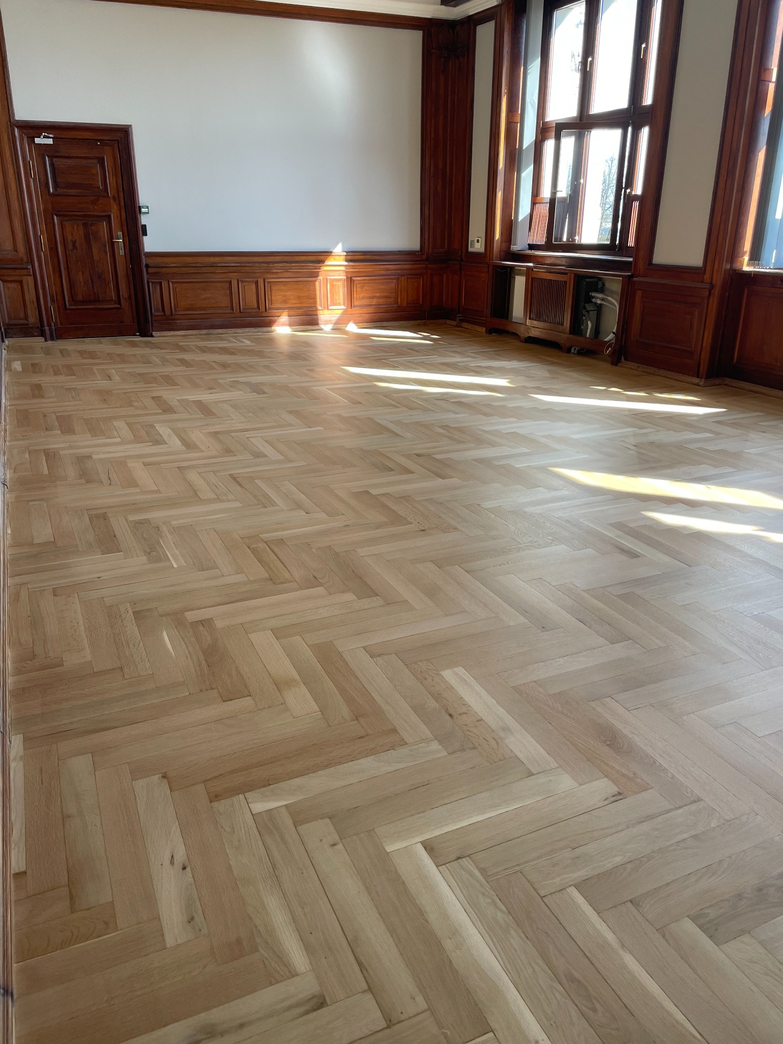 Jasny, parkiet ułożony w jodełkę w przestronnym pokoju z drewnianymi panelami ściennymi i dużymi oknami, wpadające promienie słoneczne.