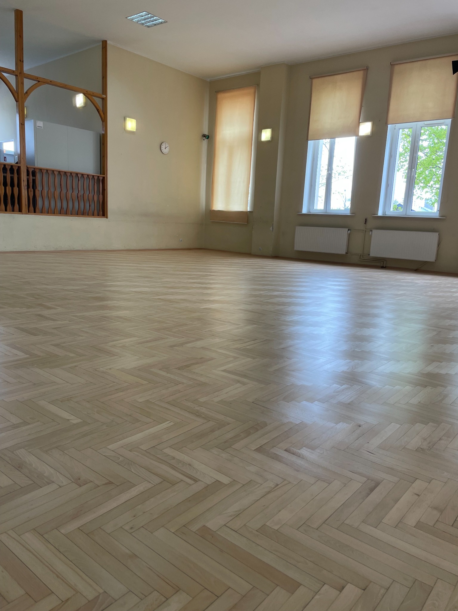 Jasny parkiet ułożony w jodełkę w przestronnym, słonecznym pokoju z drewnianą balustradą balkonu i oknami z roletami, ukazujący świeżo wykończoną podłogę.