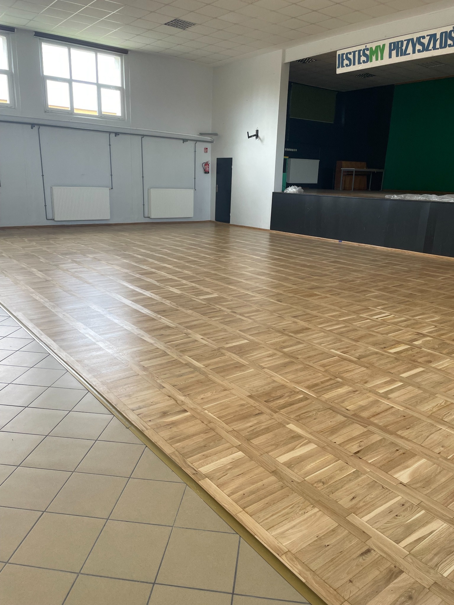 Sala gimnastyczna z nową, jasną podłogą parkietową ułożoną we wzór kwadratów. Widoczne okna i scena w tle. Parkiet łączy się z płytkami podłogowymi.