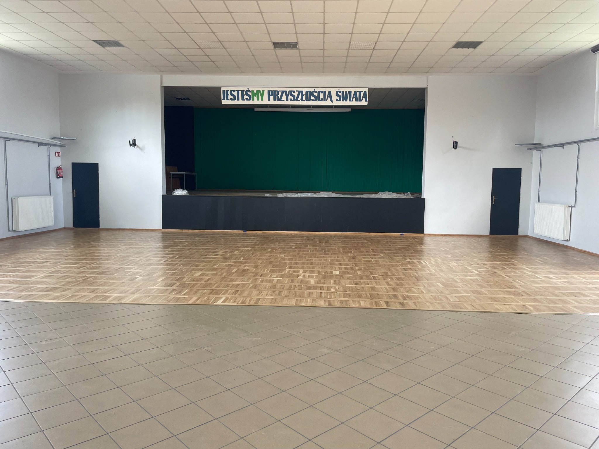 Sala z parkietem ułożonym we wzór jodełki, kontrastującym z szarymi płytkami. Scena z zielonym tłem i napisem 'Jesteśmy Przyszłością Świata' nad nią.