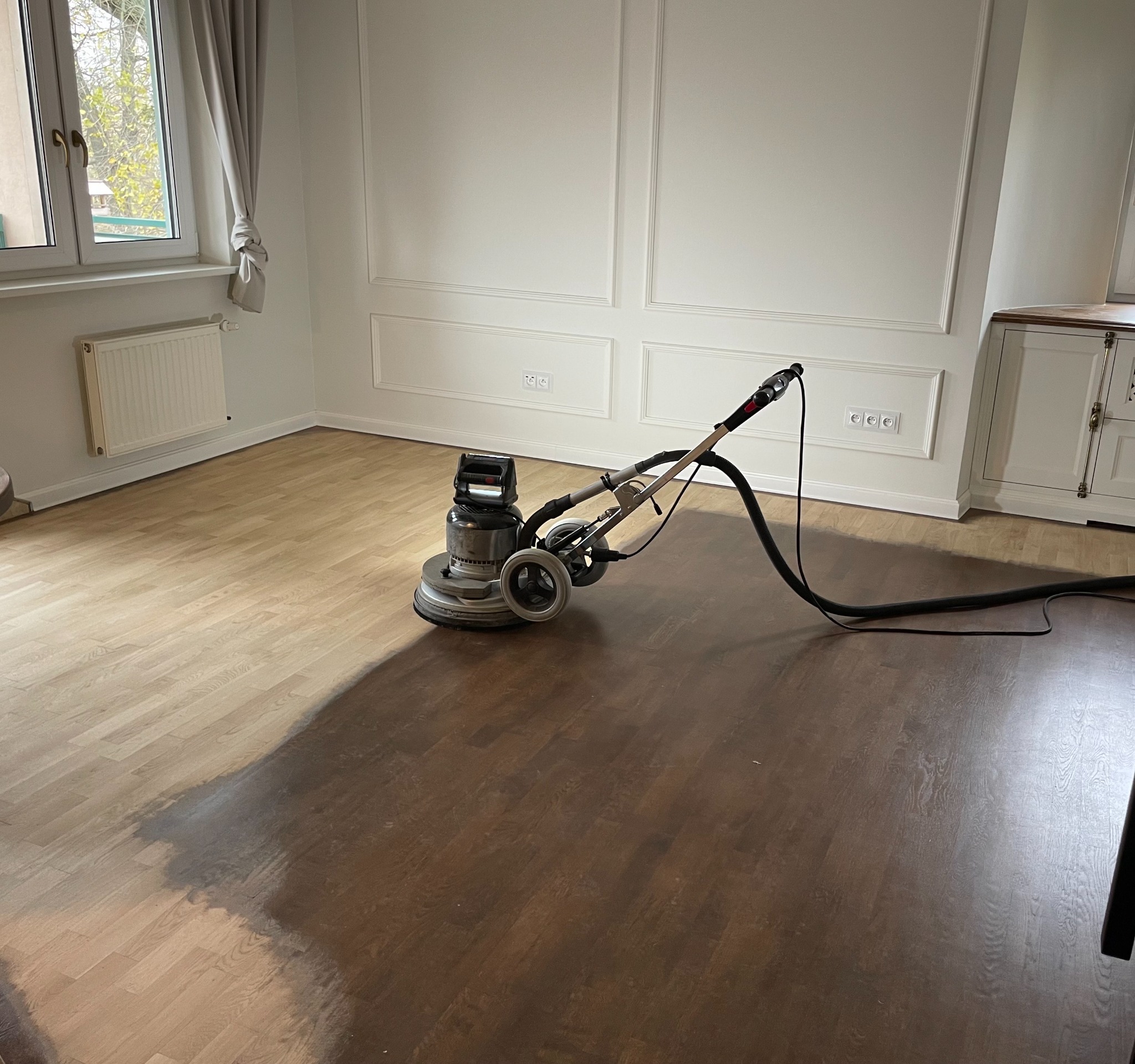 Cyklinowanie parkietu w pokoju: połowa podłogi wycyklinowana, ukazując jaśniejszy odcień drewna, maszyna do cyklinowania w trakcie pracy, eleganckie białe ściany.