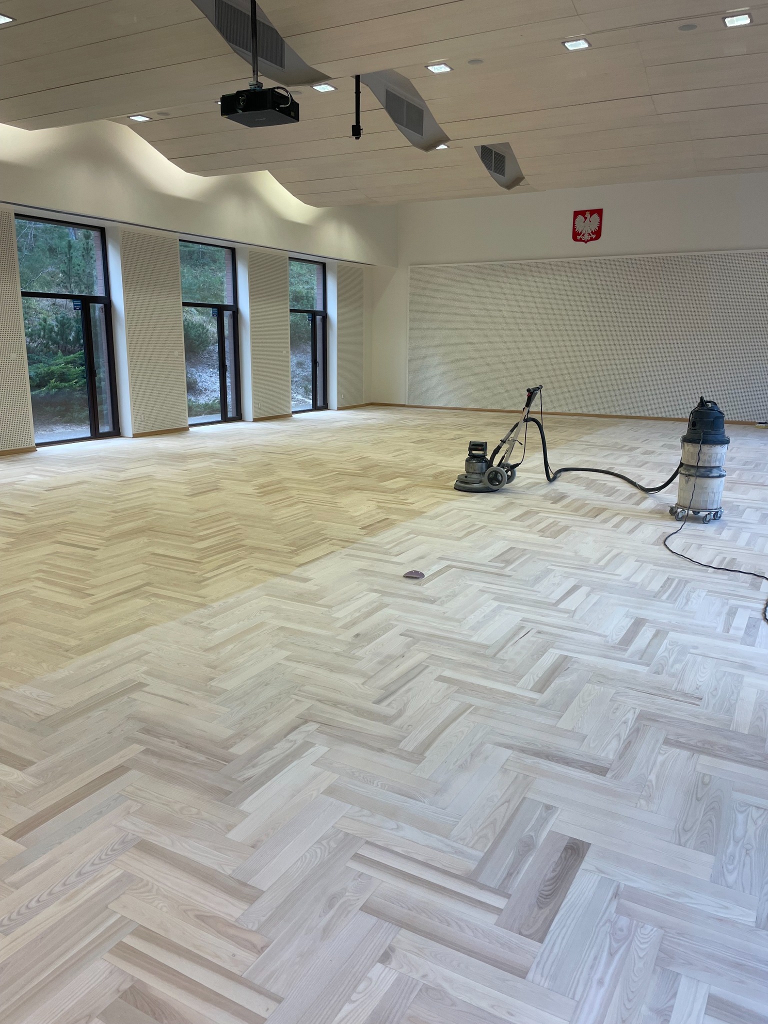 Sala z jasnym parkietem w jodełkę, częściowo wyszlifowanym, z maszyną do cyklinowania i odkurzaczem przemysłowym. W tle okna i godło Polski.