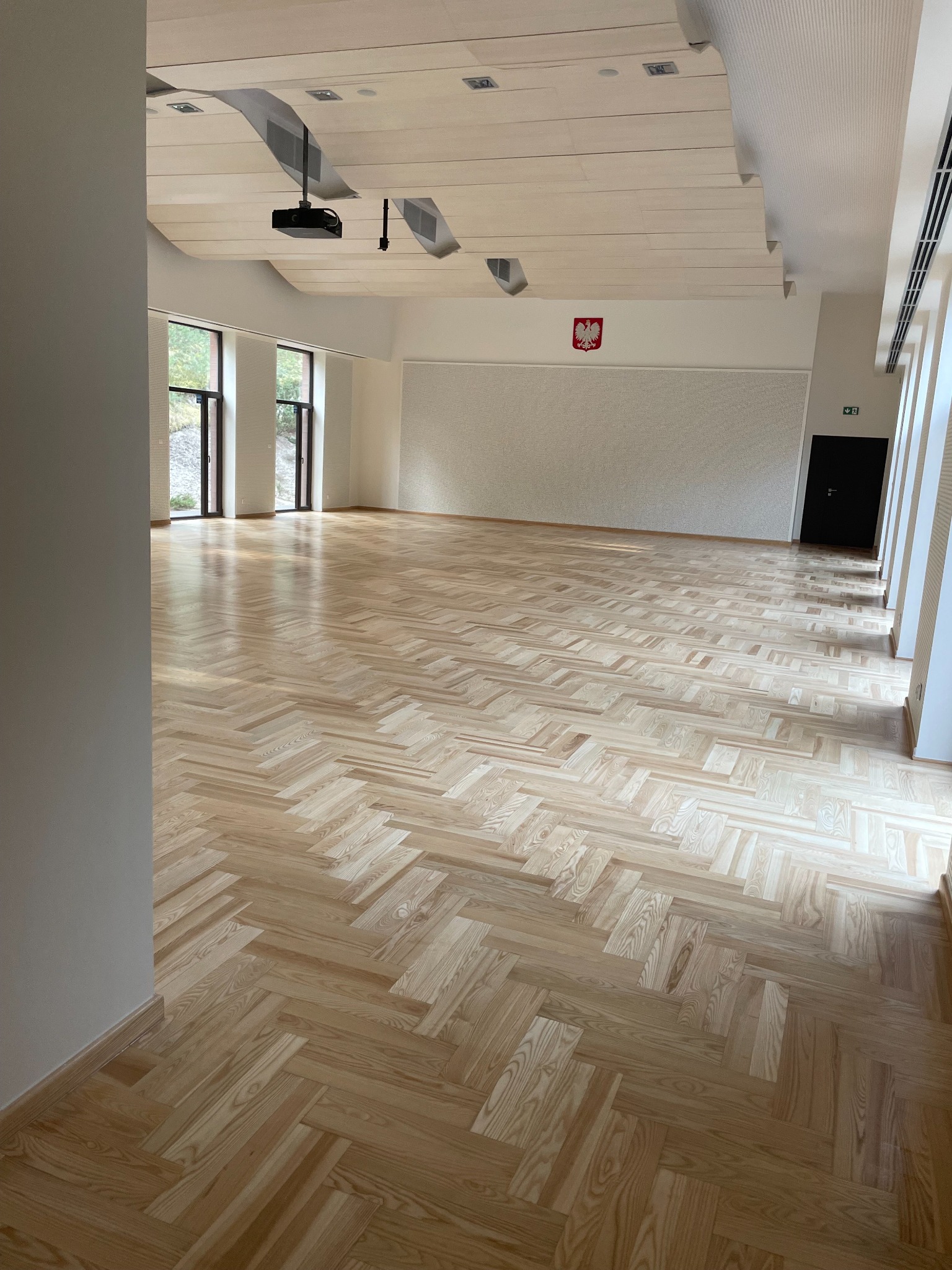 Jasny parkiet ułożony w jodełkę w przestronnym, jasnym pomieszczeniu z białymi ścianami i oknami. Na ścianie godło Polski. Widoczny projektor pod sufitem.