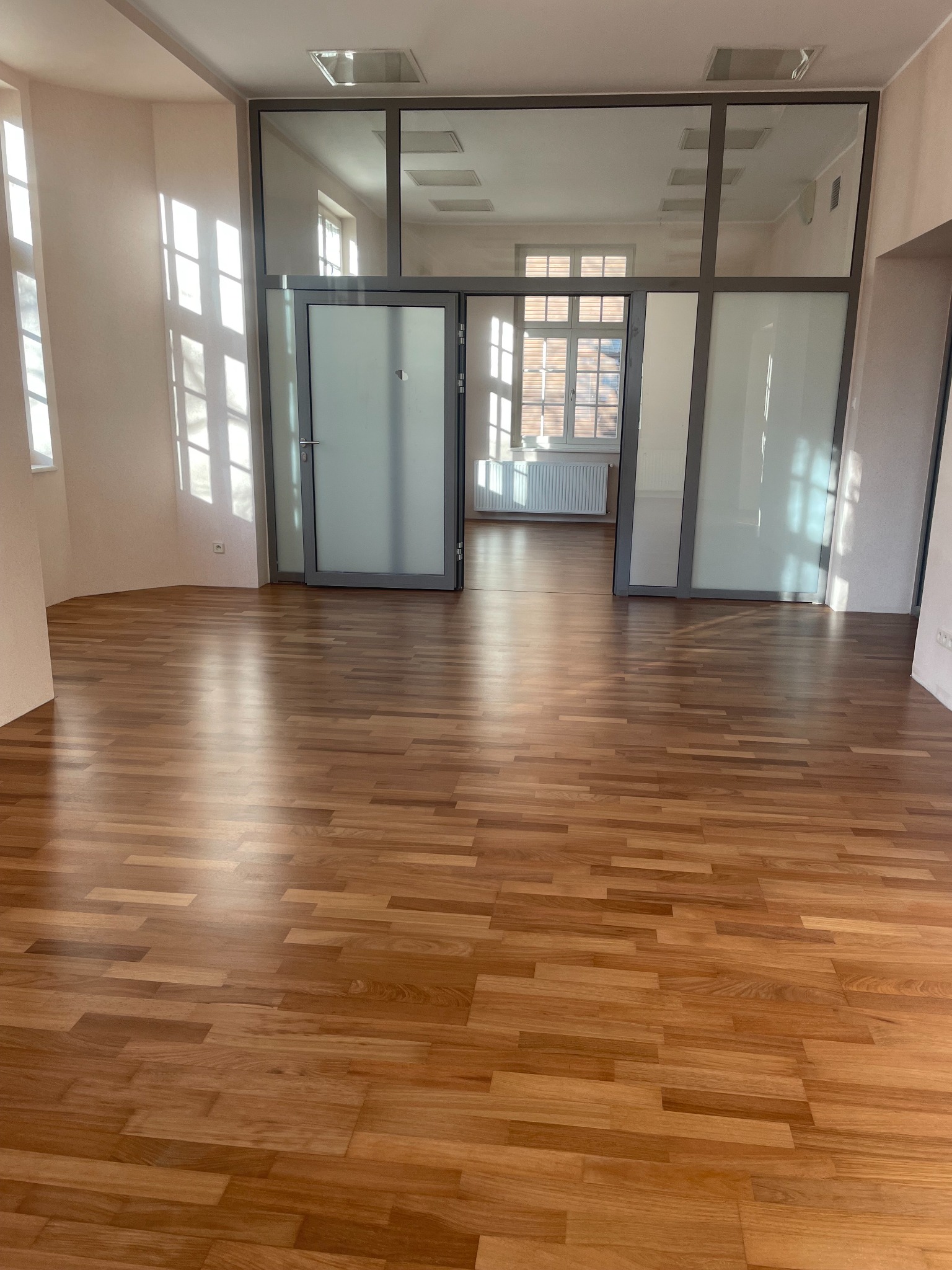Jasny parkiet w pomieszczeniu z dużymi oknami i szklanymi drzwiami, widok na kolejne pomieszczenie. Słoneczne refleksy na podłodze.