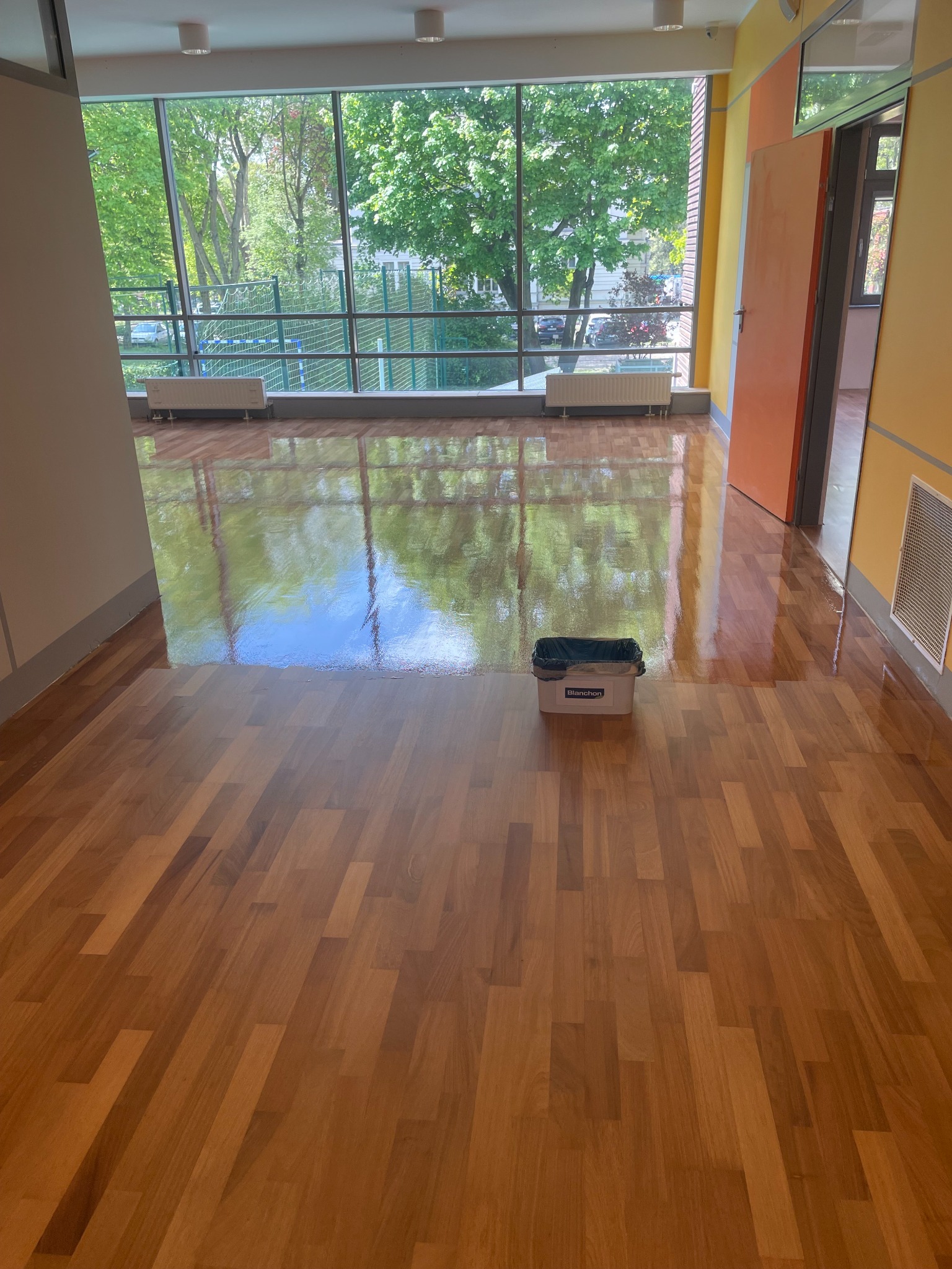 Błyszczący parkiet w jasnym pomieszczeniu z dużym oknem, widoczny pojemnik marki Blanchon. Podłoga odbija światło i zieleń z zewnątrz.