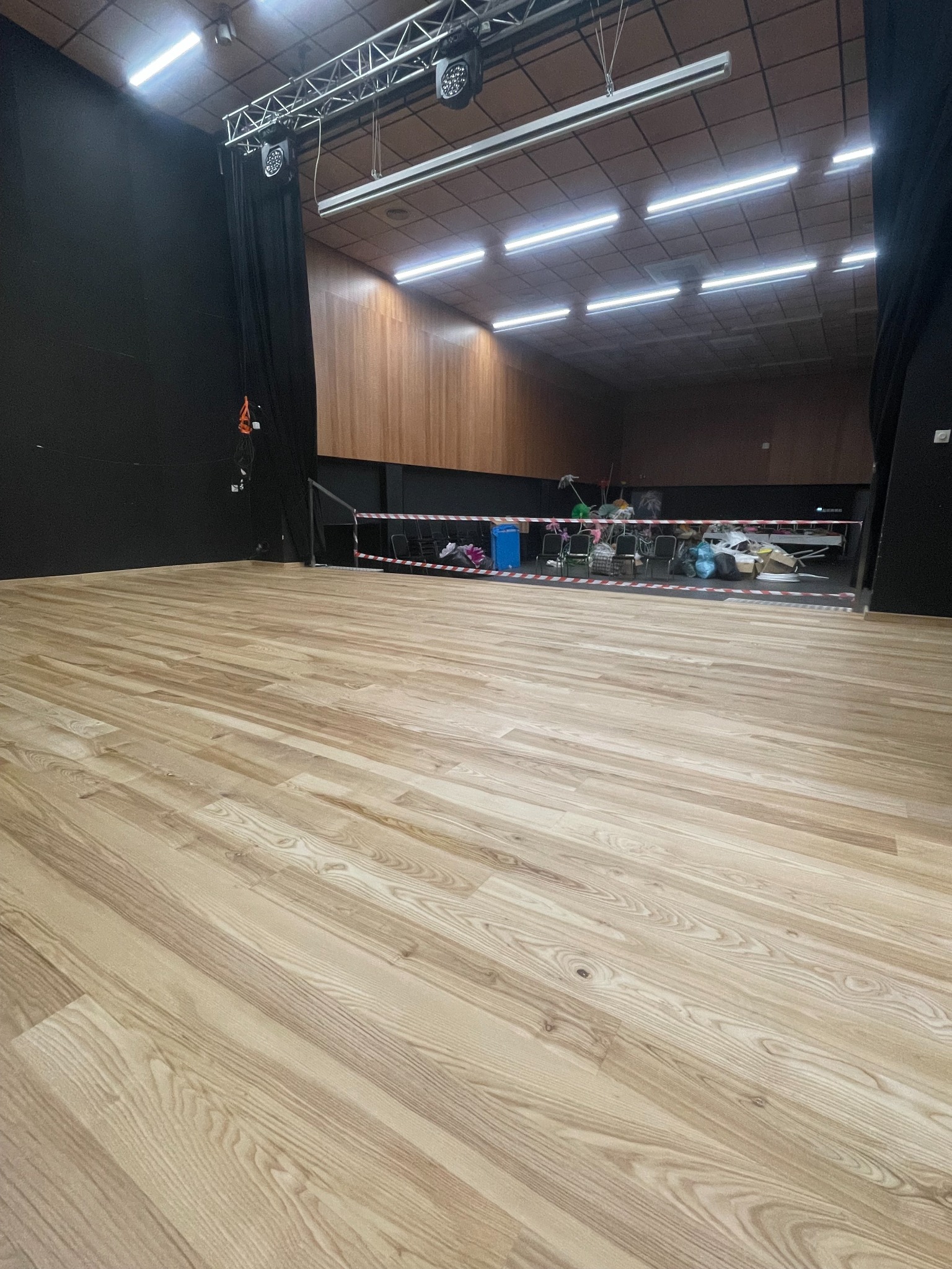 Jasne panele podłogowe w dużej sali z czarnymi zasłonami i oświetleniem sufitowym. W tle krzesła i taśma ostrzegawcza.