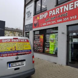 Budynek firmy BHP Partner z reklamą na fasadzie i oknach oraz oznakowany samochód dostawczy Volkswagen Caddy z numerem telefonu i adresem strony internetowej, zaparkowany przed budynkiem.