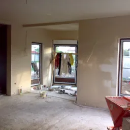 Wnętrze w trakcie prac wykończeniowych: świeżo otynkowane ściany w jasnym odcieniu, widoczne okna z suszącym się praniem na zewnątrz, na podłodze resztki materiałów budowlanych i czerwony agregat...