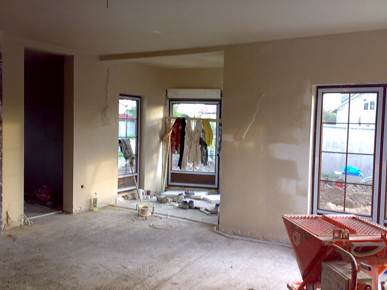 Wnętrze w trakcie prac wykończeniowych: świeżo otynkowane ściany w jasnym odcieniu, widoczne okna z suszącym się praniem na zewnątrz, na podłodze resztki materiałów budowlanych i czerwony agregat...