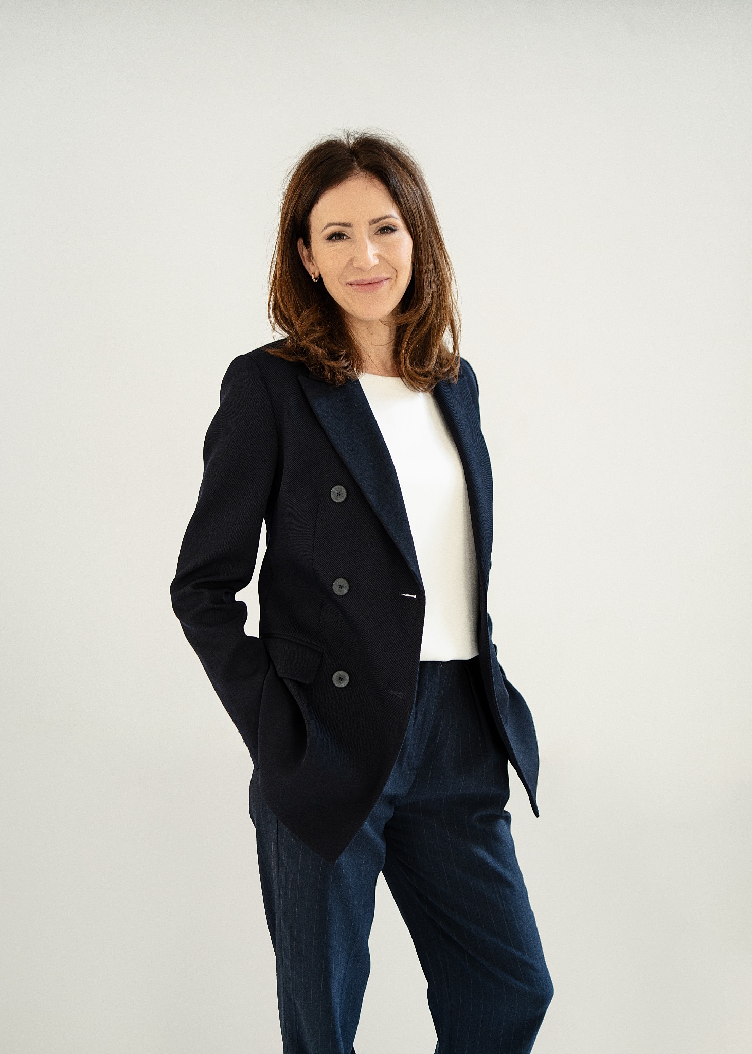 Portret uśmiechniętej kobiety w granatowym, dwurzędowym garniturze w prążki i białej bluzce na tle jasnego studia.