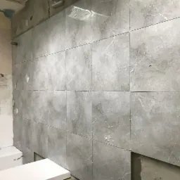 Ściana łazienki w trakcie układania szarych płytek imitujących marmur, z widocznymi krzyżykami dystansowymi i fragmentem surowej ściany obok.