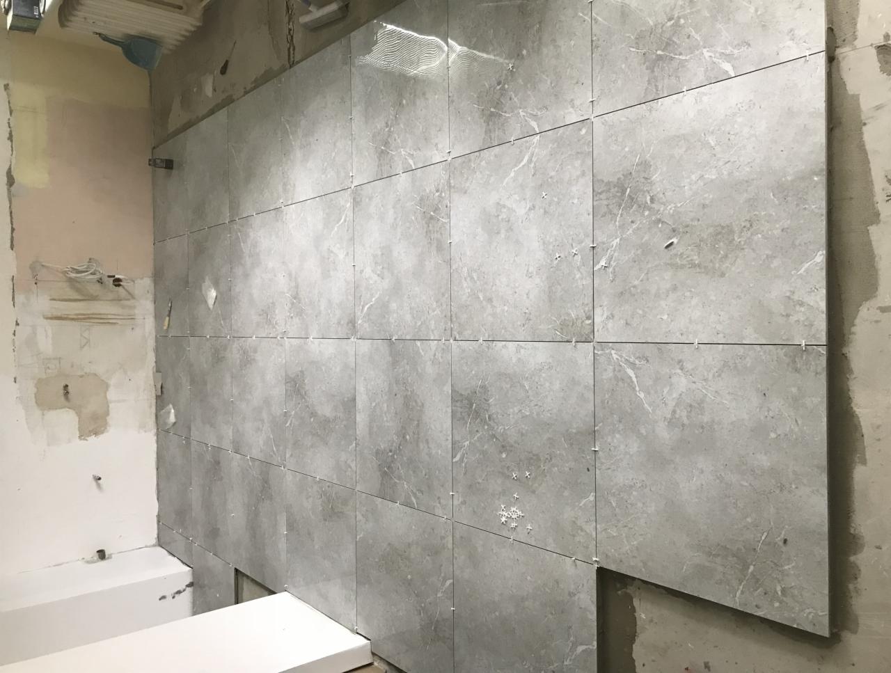 Ściana łazienki w trakcie układania szarych płytek imitujących marmur, z widocznymi krzyżykami dystansowymi i fragmentem surowej ściany obok.