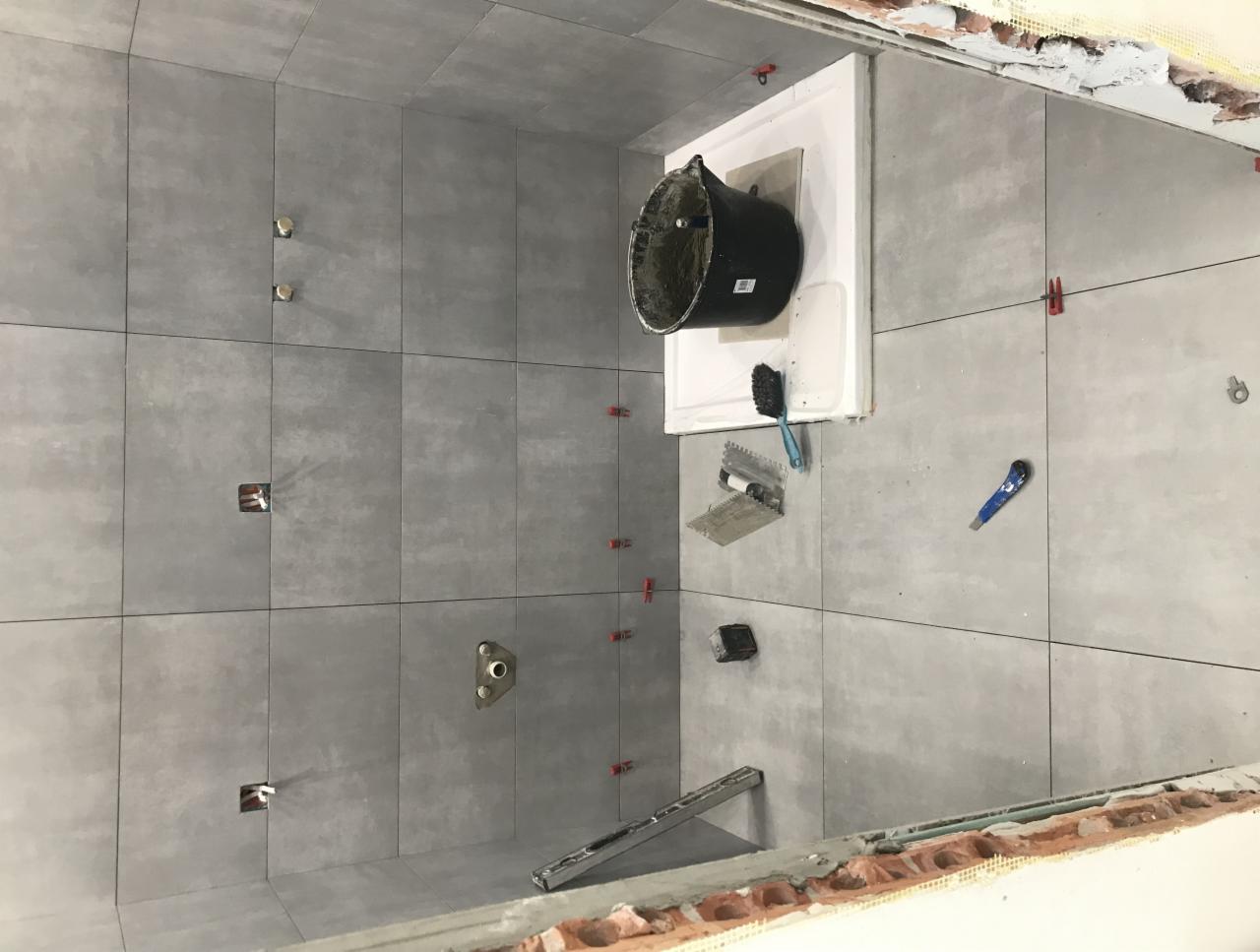 Wnętrze łazienki w trakcie remontu, widoczne szare płytki na ścianach, czarne wiadro na białej obudowie, narzędzia do układania płytek i czerwone klipsy poziomujące.