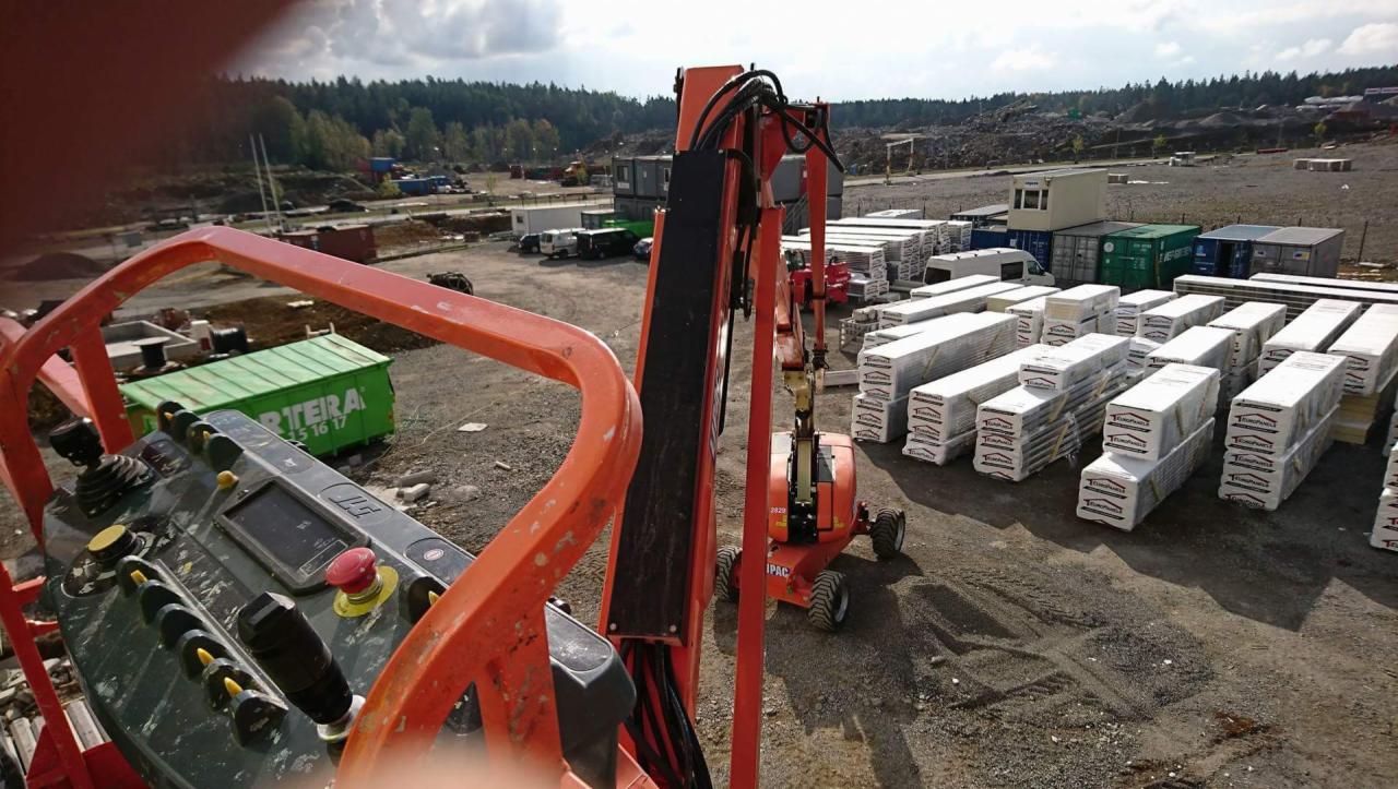 Widok z podnośnika koszowego na plac budowy z przygotowanymi elementami konstrukcyjnymi, prawdopodobnie panelami warstwowymi, składowanymi w pakietach, kontenery biurowe w tle.