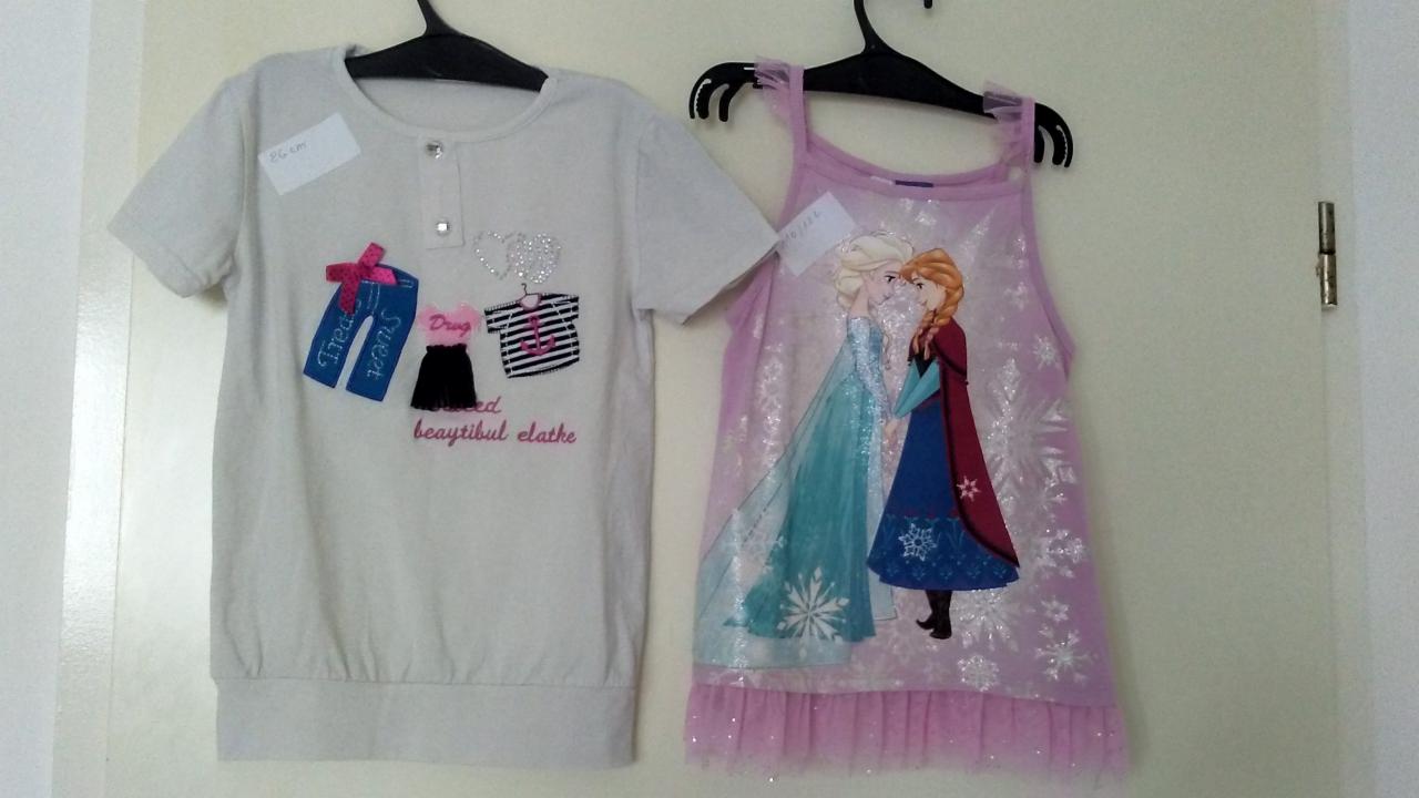 Dwie dziecięce bluzki na wieszakach: biała z aplikacjami ubrań i napisami oraz liliowa z grafiką Elsy i Anny z 'Krainy Lodu', obie z widocznymi metkami.