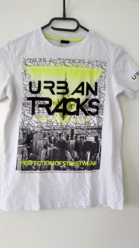 Biała koszulka z nadrukiem miejskiego krajobrazu i napisem 'URBAN TRACKS' na wieszaku.