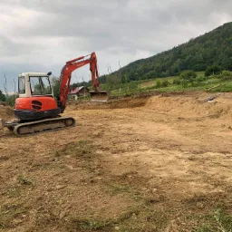 Pomarańczowa minikoparka Kubota podczas wykopywania fundamentów pod budynek na tle zalesionego wzgórza i pochmurnego nieba.