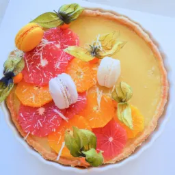 Tarta z kremem cytrynowym, udekorowana plastrami grejpfruta i pomarańczy, makaronikami oraz miechunką peruwiańską w białej formie do pieczenia.