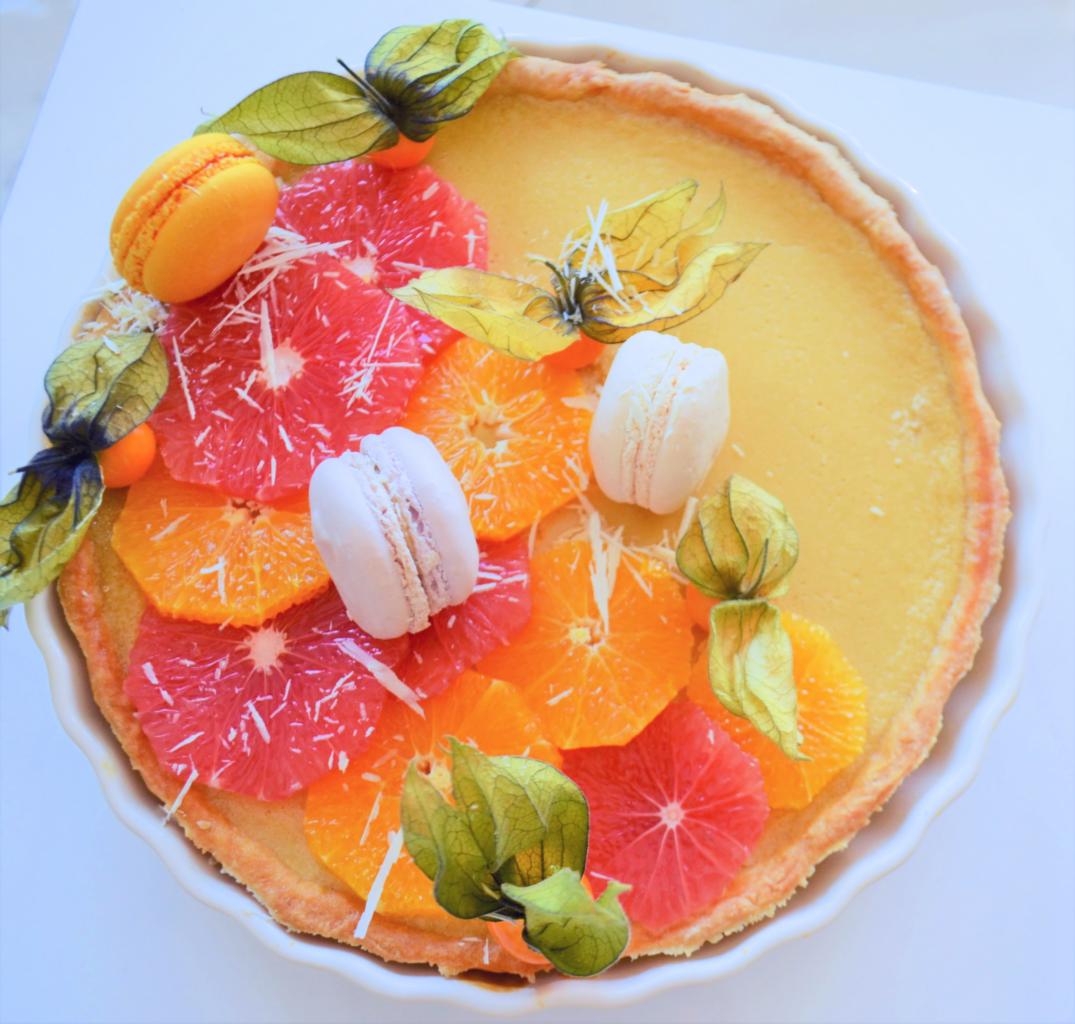 Tarta z kremem cytrynowym, udekorowana plastrami grejpfruta i pomarańczy, makaronikami oraz miechunką peruwiańską w białej formie do pieczenia.