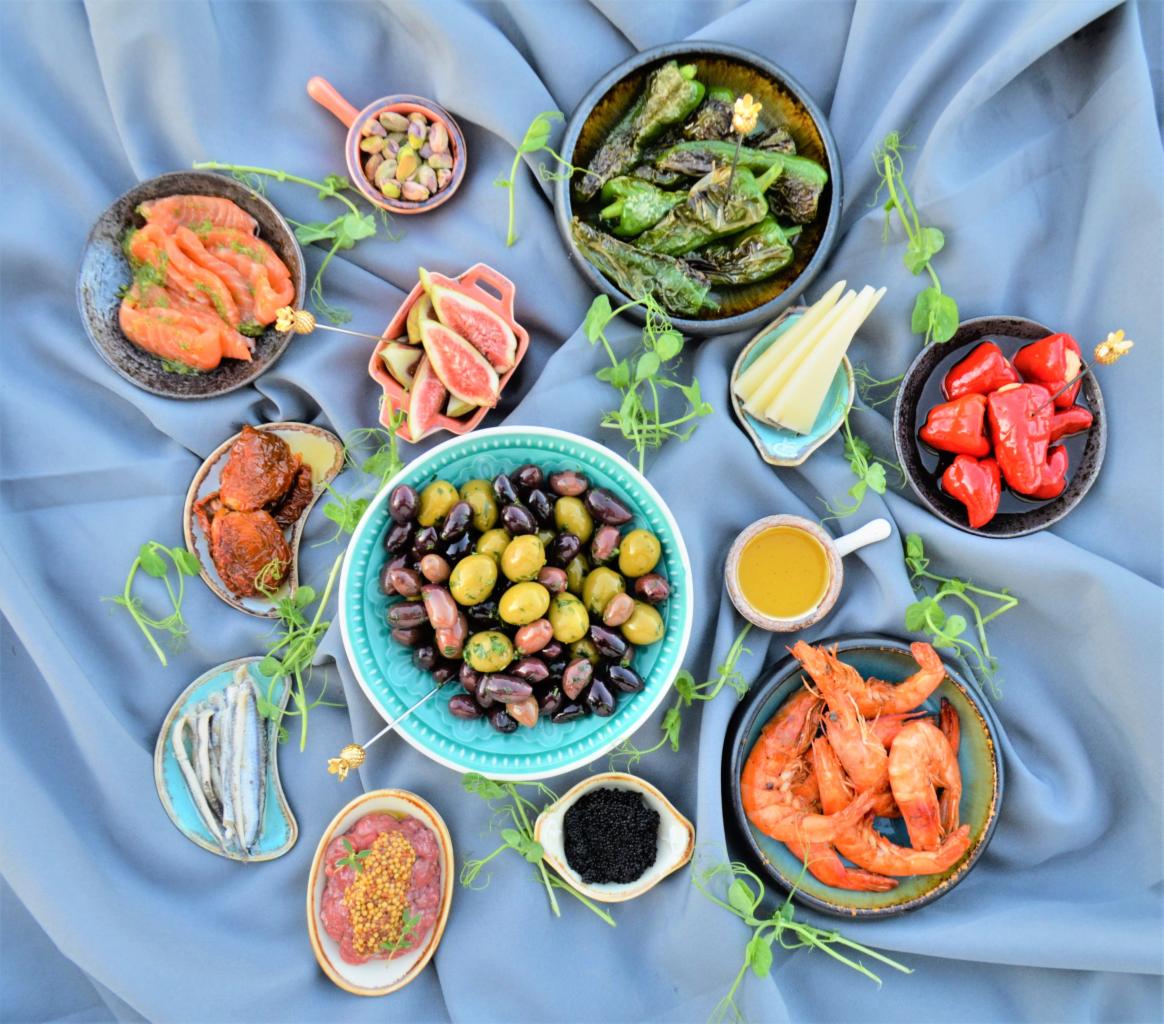 Elegancki układ różnorodnych przekąsek w małych naczyniach na pogniecionym, błękitnym tle, z dekoracyjnymi pędami roślin. Widoczne są m.in. oliwki, krewetki, ryby, sery, papryczki i pistacje.