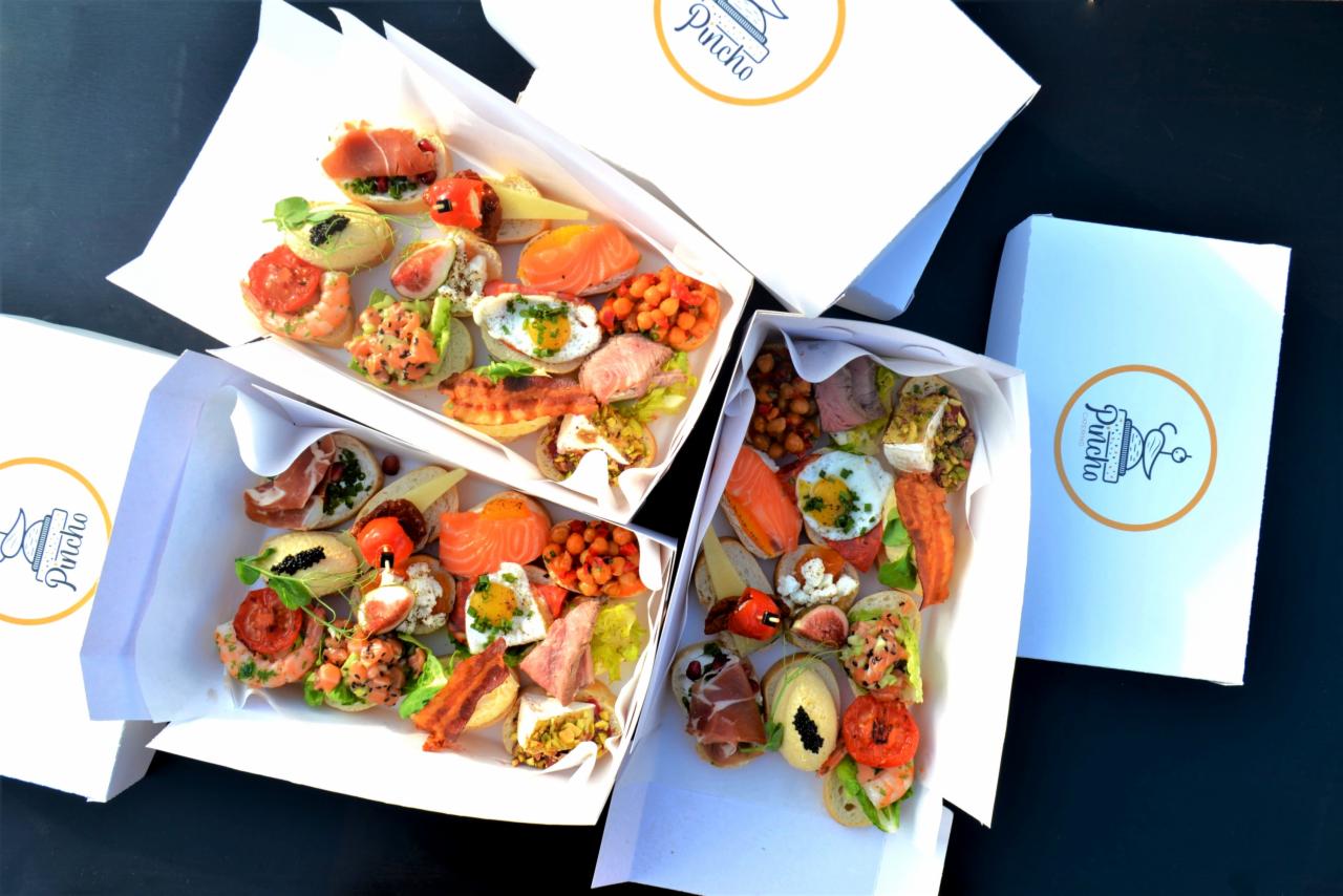 Elegancko zapakowane, różnorodne kanapki bankietowe w białych pudełkach z logo na ciemnym tle, widoczne różne rodzaje pieczywa i dodatków, takich jak łosoś, jajka przepiórcze, szynka parmeńska...