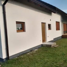 Ocieplona elewacja domu jednorodzinnego w Polkowicach, widoczne brązowe okna i drzwi, oraz szara podmurówka i brązowe rynny.