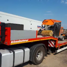 Ciężarówka niskopodwoziowa marki Kässbohrer przewozi pomarańczową zamiatarkę drogową H99 4WD Optimus na tle białej naczepy i błękitnego nieba.