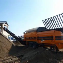 Pomarańczowa przesiewarka mobilna Doppstadt SM 518 z podajnikiem taśmowym usypuje pryzmę ziemi na placu budowy, w tle budynek z jasną elewacją i błękitne niebo.