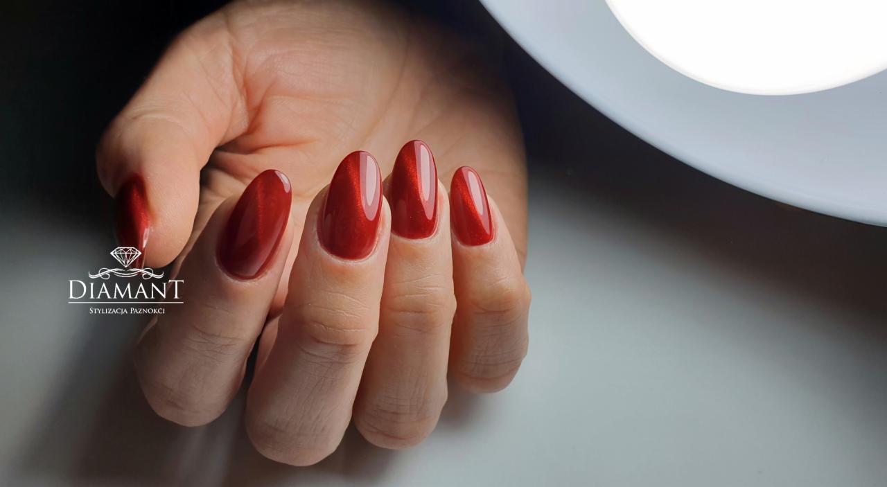 Dłoń z błyszczącymi, czerwonymi paznokciami w kształcie migdała, z efektem 'kocie oko', nałożonym na naturalną płytkę, w tle lampa UV i logo salonu.