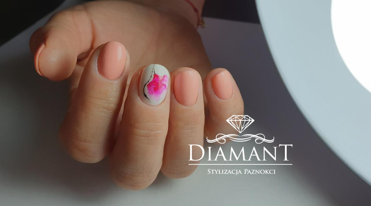 Dłoń z paznokciami pomalowanymi na pastelowy brzoskwiniowy kolor, jeden paznokieć z białym tłem i malowanym różowym kwiatem z czarnymi konturami, widoczne logo firmy w prawym dolnym rogu.