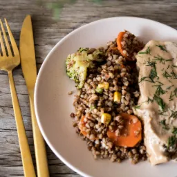 Porcja kaszy gryczanej z marchewką, brokułem i sosem na talerzu, obok złote sztućce na drewnianym tle.