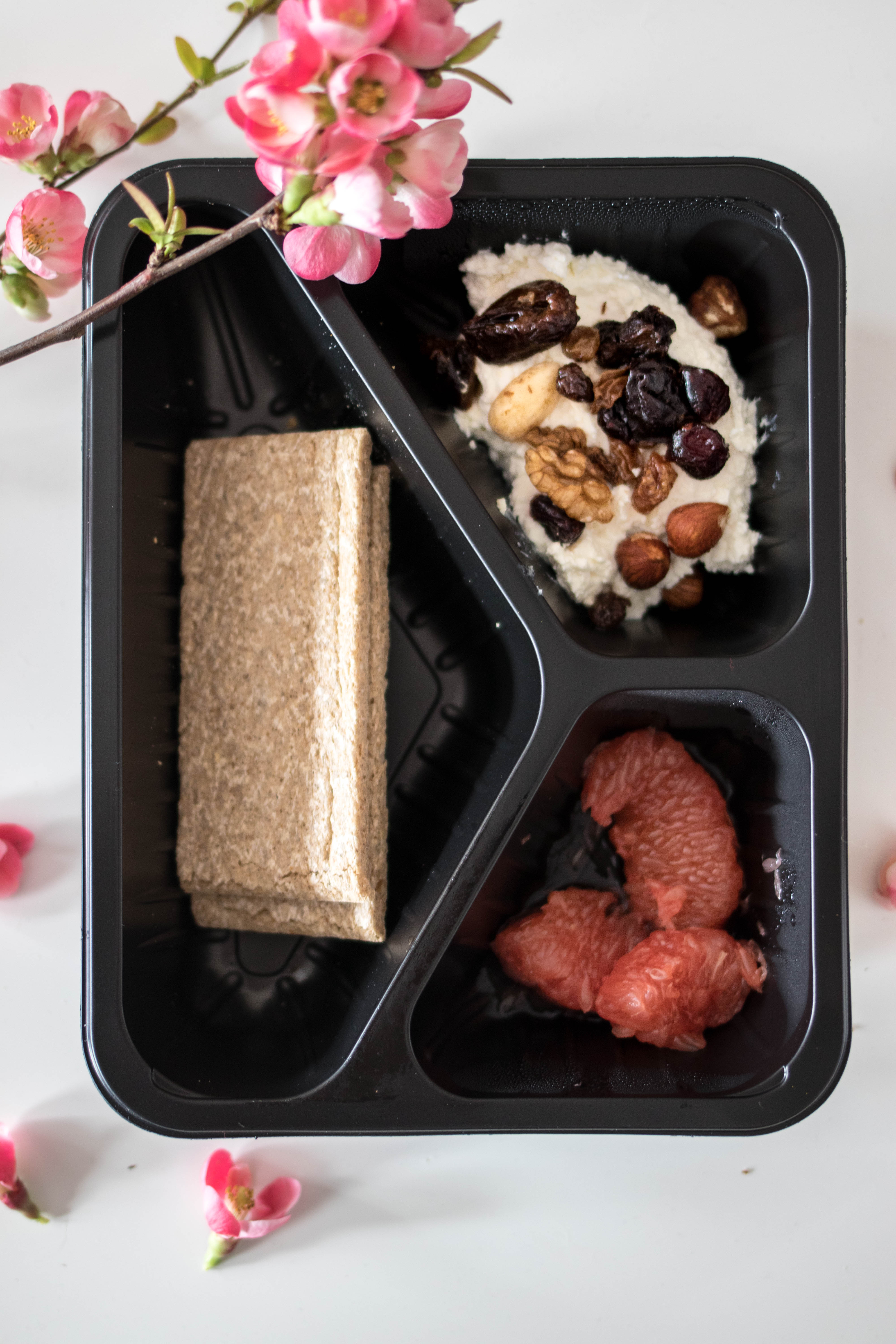 Posiłek w czarnym pudełku z trzema przegródkami: pieczywo chrupkie, twarożek z orzechami i suszonymi owocami, cząstki grejpfruta. Dekoracja z kwitnącej gałązki.