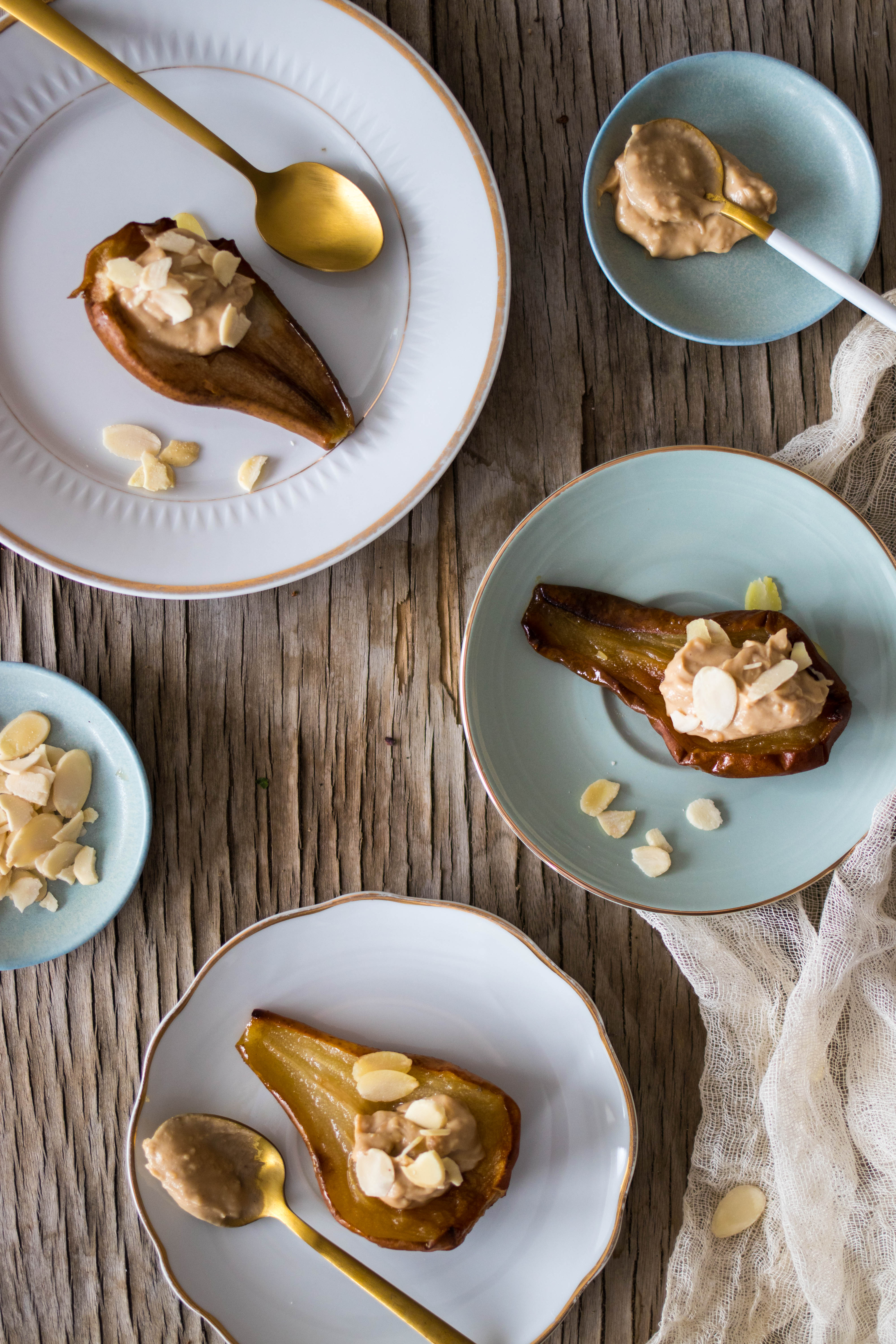 Karmelizowana połówka gruszki z kremowym nadzieniem i płatkami migdałów na talerzach z ozdobnymi brzegami, ułożonych na surowym drewnianym stole z lnianą ściereczką. Złote łyżki obok.