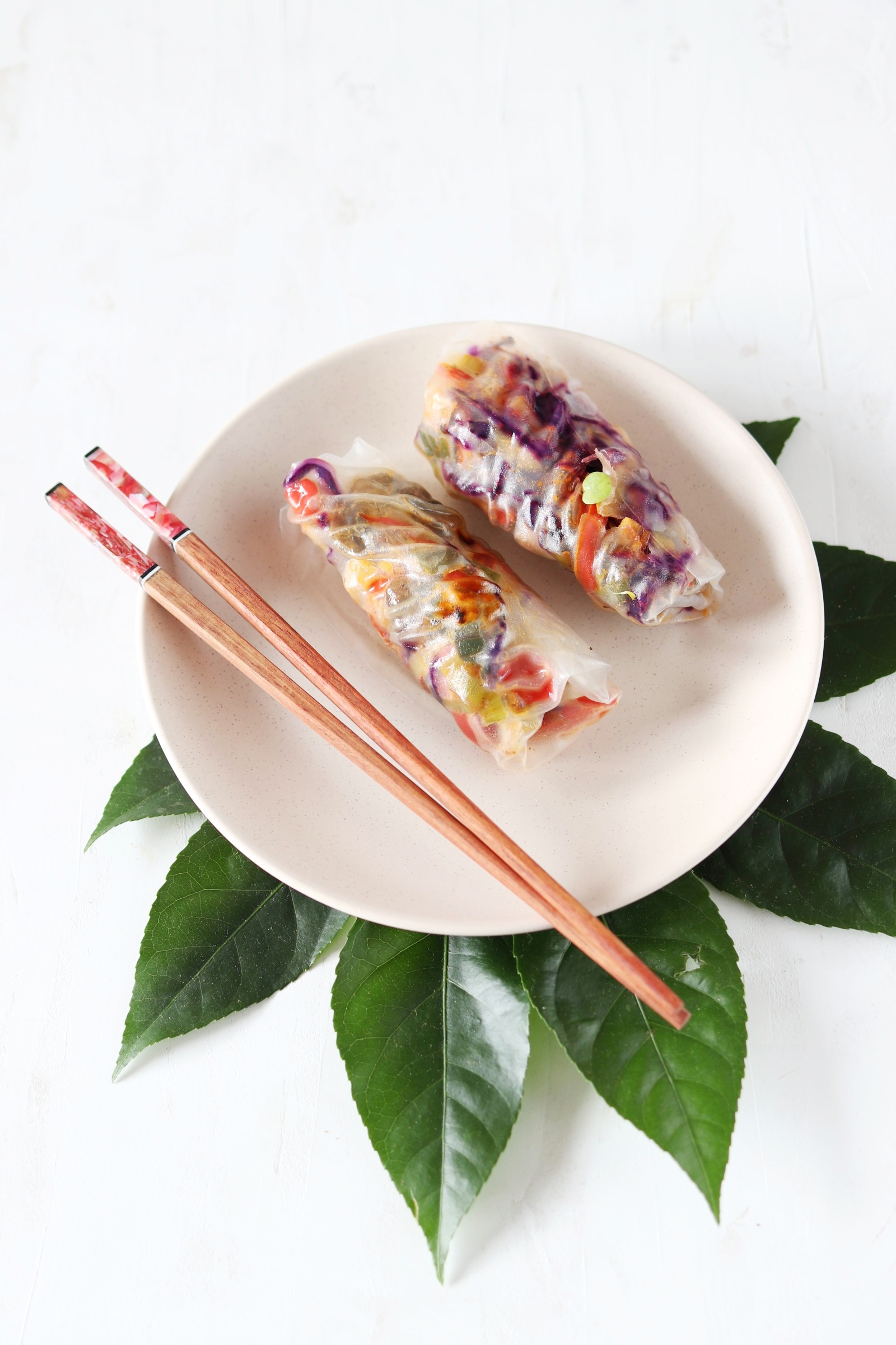 Dwa przezroczyste sajgonki wypełnione kolorowymi warzywami, ułożone na kremowym talerzu z pałeczkami i zielonymi liśćmi na białym tle.