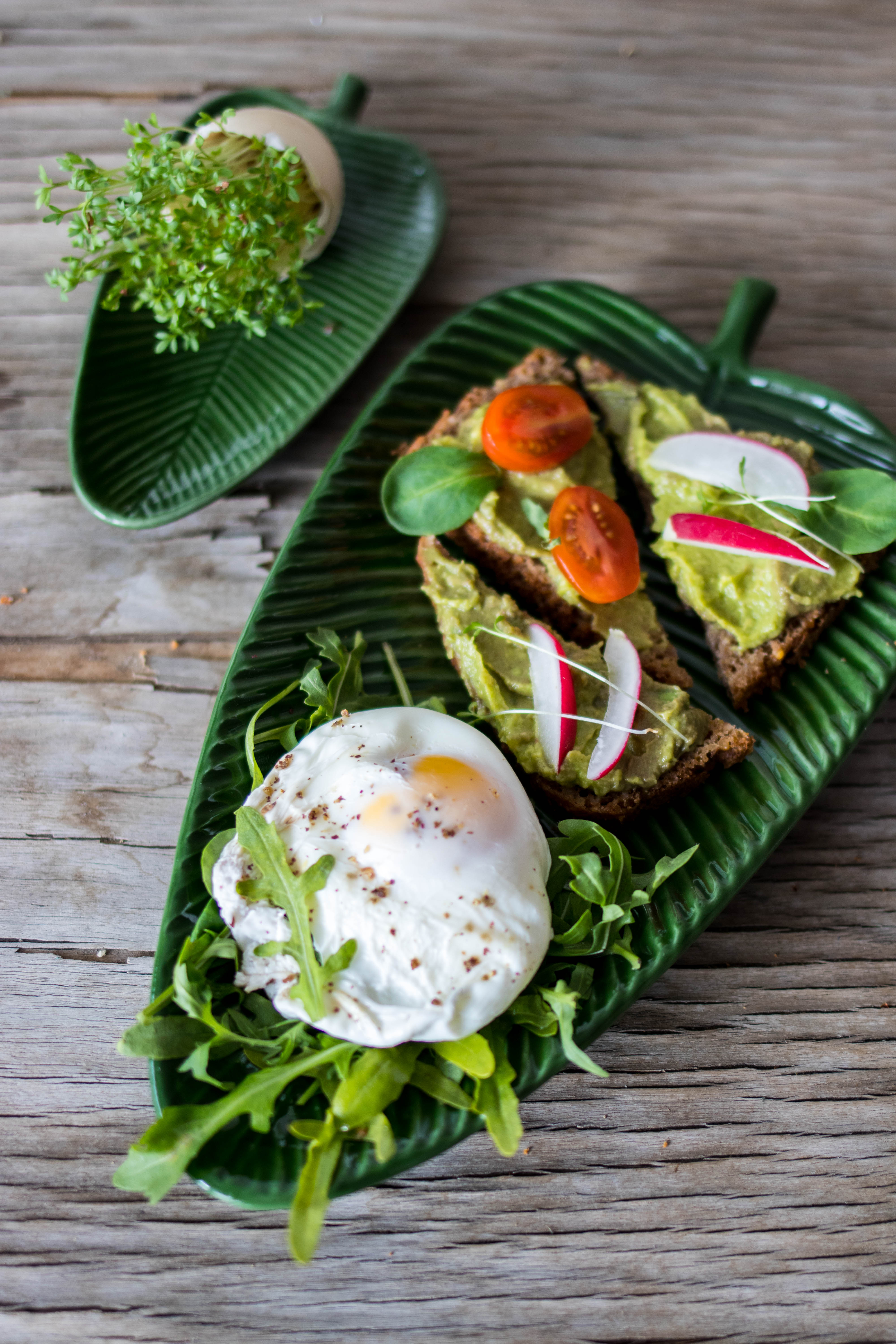 Dwa zielone talerze w kształcie liści bananowca na drewnianym stole: na jednym sadzonka rzeżuchy w skorupce jajka, na drugim kanapki z awokado, rzodkiewką i pomidorem oraz jajko sadzone na rukoli.