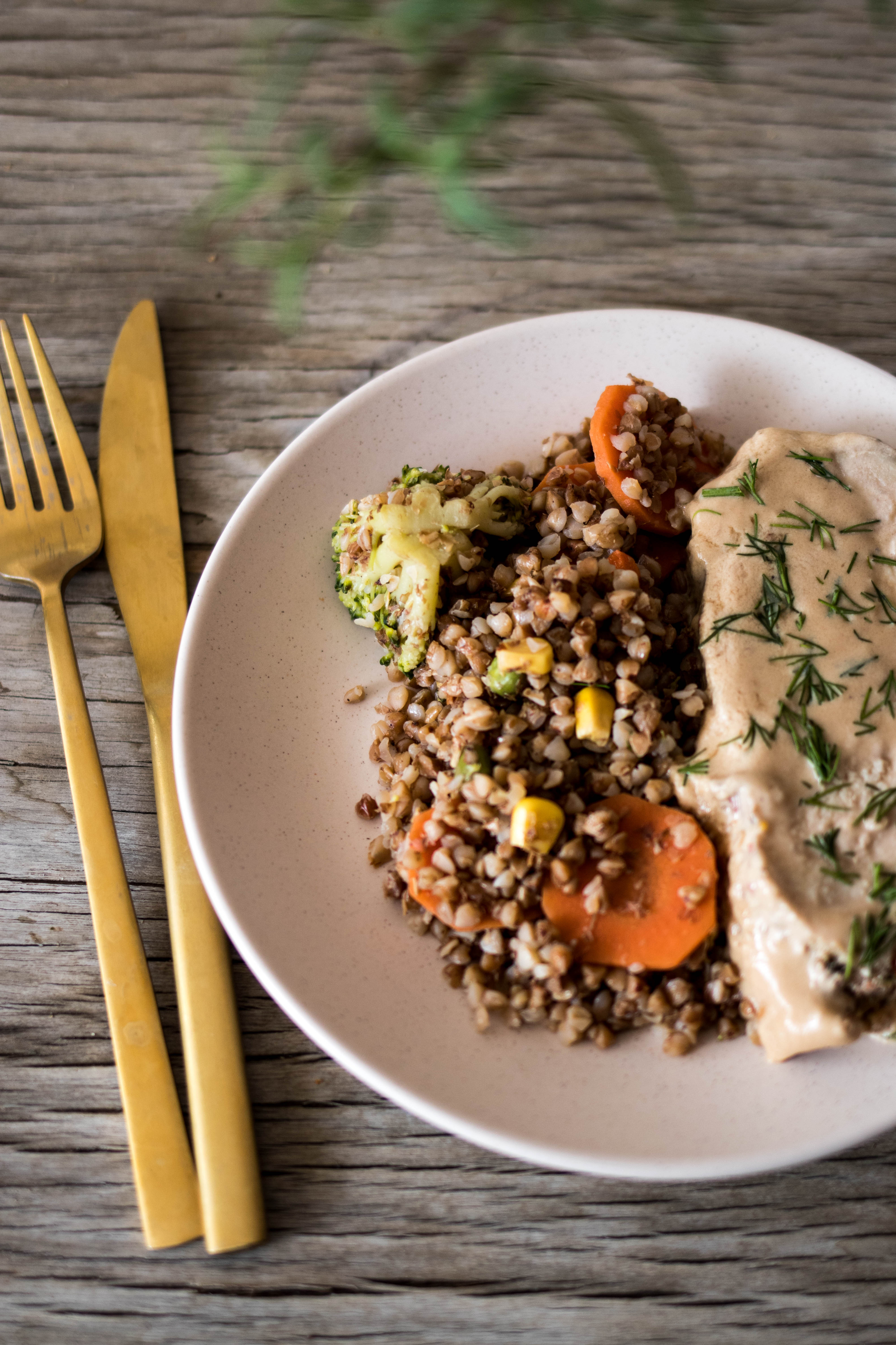 Porcja kaszy gryczanej z marchewką, brokułem i sosem na talerzu, obok złote sztućce na drewnianym tle.
