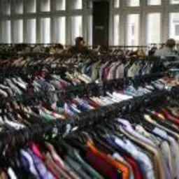 Rzędy wieszaków z używanymi koszulami w sklepie, widoczne osoby w tle, oświetlone przez duże okna.