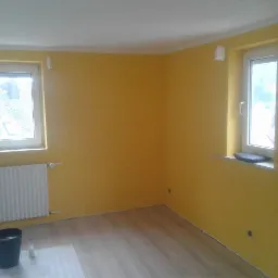Pokój po odświeżeniu, ściany pomalowane na żółto, widoczne dwa okna, grzejnik, wiadro z farbą i akcesoria malarskie na podłodze zabezpieczonej folią.