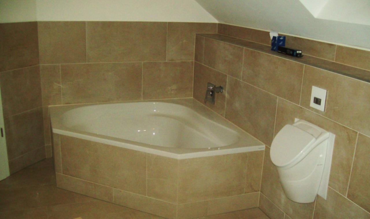 Narożna wanna obudowana beżowymi płytkami w łazience na poddaszu, z widoczną armaturą, podwieszanym bidetem i półką z dekoracyjnym modelem pociągu.