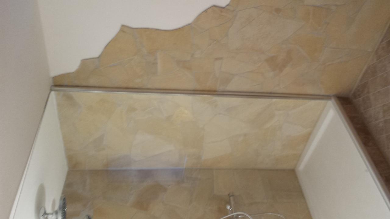 Beżowe płytki imitujące kamień na ścianach i suficie łazienki, oddzielone cienką, srebrną listwą, z widoczną armaturą prysznicową i fragmentem białej ściany.