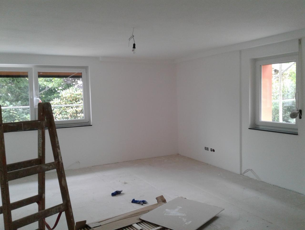 Pokój w trakcie malowania na biało z drewnianą drabiną w lewym rogu, zabezpieczona podłoga i wisząca żarówka.