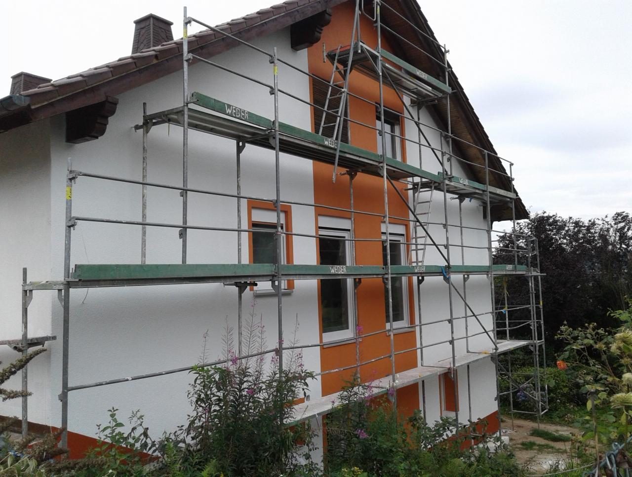 Dom w trakcie malowania elewacji, widoczne rusztowanie z zielonymi elementami, część ściany w kolorze pomarańczowym, druga część biała.