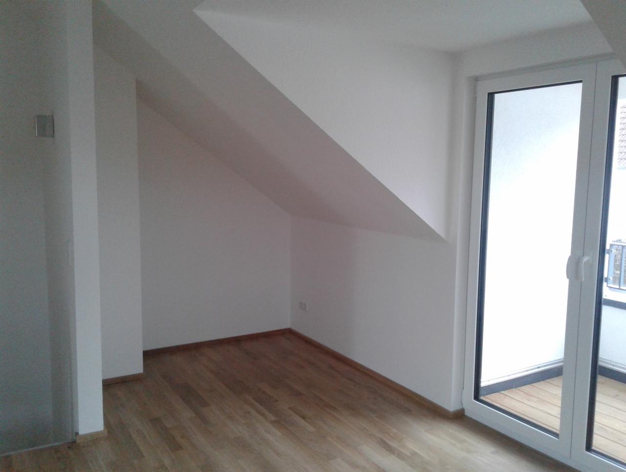 Puste, białe poddasze z drewnianą podłogą i wyjściem na balkon przez przeszklone drzwi balkonowe.