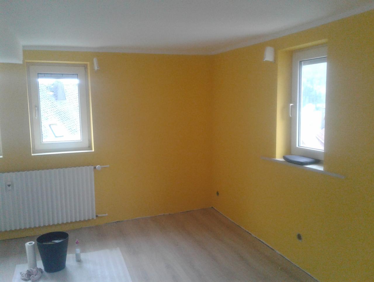 Pokój po odświeżeniu, ściany pomalowane na żółto, widoczne dwa okna, grzejnik, wiadro z farbą i akcesoria malarskie na podłodze zabezpieczonej folią.
