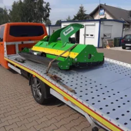 Pomarańczowy bus z platformą transportową przewozi zielono-żółtą kosiarkę marki Bomet na tle placu z kostki brukowej i budynków.